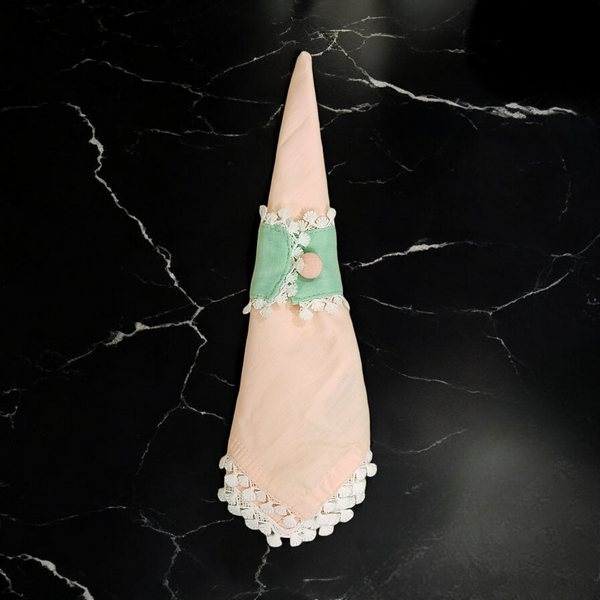 The Lotus Buti dining napkin and napkin rings featuring a peach fabric napkin with white lace and a green napkin ring